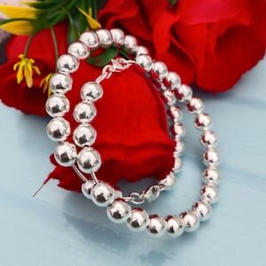 May include: Two silver beaded bracelets with a polished finish. The bracelets are composed of round, evenly sized beads. The jewelry is displayed on a red surface with a red rose and yellow flowers in the background.