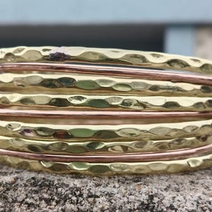 Puede incluir: Una pila de brazaletes dorados y cobrizos martillados. Los brazaletes dorados tienen una superficie texturizada y con hoyuelos, mientras que los brazaletes de cobre son lisos. Los brazaletes están apilados sobre una superficie rugosa y texturizada.