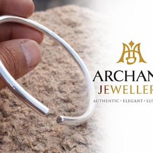 Puede incluir: Un brazalete de plata con un diseño abierto. El brazalete se sostiene contra una superficie texturizada de color marrón claro. El logotipo de Archana Jewellers es visible en el fondo, con las palabras "Auténtico, Elegante, Lujoso".
