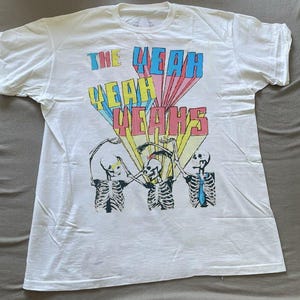 Può includere: T-shirt bianca con la scritta "THE YEAH YEAH YEAH'S" in blu, giallo e rosa, in stile retrò. Il design include tre figure di scheletri. La maglietta è realizzata in un materiale morbido.
