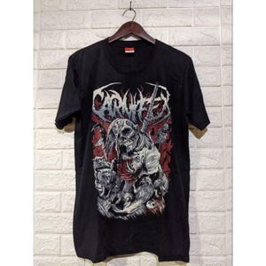 May include: Black t-shirt featuring a graphic of a skull with long hair, holding a weapon, and surrounded by other figures. The word "CARNIFEX" is printed above the skull in a stylized font. The design has red and white accents.