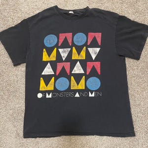 Op de afbeelding: Zwart t-shirt met een kleurrijk geometrisch ontwerp met rode, blauwe, gele en witte vormen. De tekst "OF MONSTERS AND MEN" staat in het wit onderaan het t-shirt.