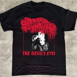 May include: Black t-shirt with a red and white graphic. The graphic features a person wearing a mask, with red abstract shapes above and the text "THE DEVIL'S EYES" in red.