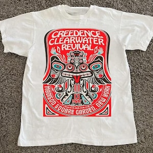May include: White t-shirt featuring a vibrant graphic design for the band Creedence Clearwater Revival. The design includes the band's name, a stylized illustration, and the text "Madison Square Garden, New York, 13 May 1970".