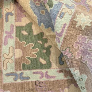 K&ouml;nnte beinhalten: Nahaufnahme eines gemusterten Teppichs in Beige-, Gr&uuml;n-, Lila- und Blaut&ouml;nen. Der Teppich zeigt ein geometrisches Design mit floralen und abstrakten Formen. Der Text "CARPETS CITY" ist sichtbar.