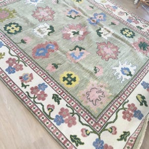 May include: A large, rectangular rug with a light green background and a floral pattern. The rug features pink, blue, and white flowers, with a cream-coloured border adorned with more flowers and a red trim. The rug is made of a soft material.