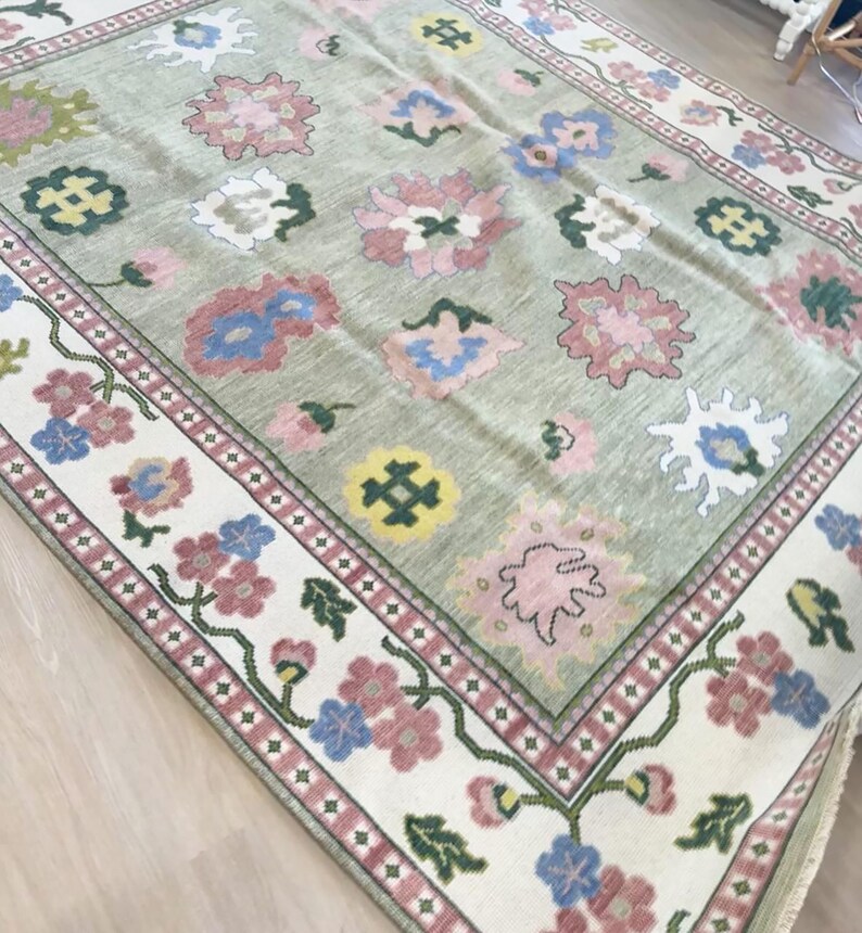 May include: A large, rectangular rug with a light green background and a floral pattern. The rug features pink, blue, and white flowers, with a cream-coloured border adorned with more flowers and a red trim. The rug is made of a soft material.