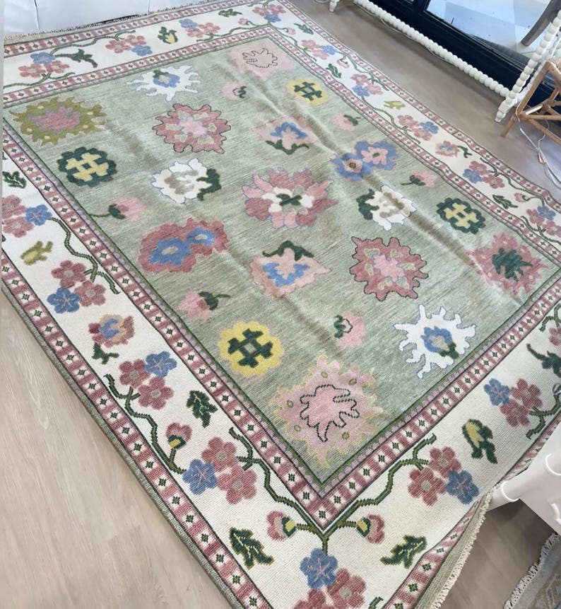 May include: A rectangular rug with a light green central field and a floral pattern. The rug has a cream border with pink, blue, and green floral designs. The central field features large, stylized flowers in pink, blue, and white.