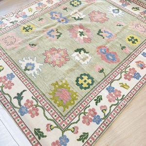 May include: A rectangular rug with a light green central field and a floral pattern. The rug has a border with pink, blue, and green flowers on a cream background. The centre has large, colourful floral designs.