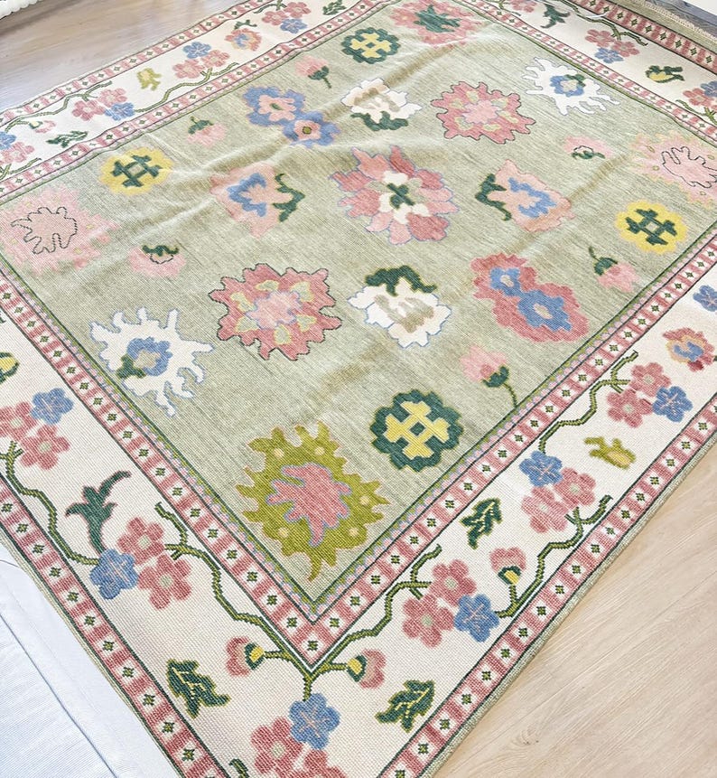 May include: A rectangular rug with a light green central field and a floral pattern. The rug has a border with pink, blue, and green flowers on a cream background. The centre has large, colourful floral designs.