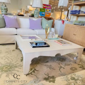 May include: A white square coffee table with a decorative edge, holding books and decorative items. The table sits on a floral patterned rug. A white sofa with purple pillows is in the background. The text "CARPETS CITY" is visible.