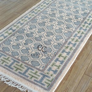 Handmade wool runner rug featuring a geometric lattice design in soft beige, blue, and green tones with tribal border details. This long hallway rug adds timeless style to entryways, kitchens, or bedside décor. Durable handwoven construction with fringe edges enhances farmhouse, boho, and modern interiors. Neutral yet colorful design makes it versatile for layering or as a statement runner. Perfect handmade rug for hallways, narrow spaces, and unique home décor accents.