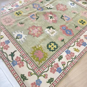 May include: A rectangular rug with a light green central field and a cream border. The rug features floral patterns in pink, blue, and yellow. The border has a repeating floral design with pink and green accents. The rug's dimensions are not visible.