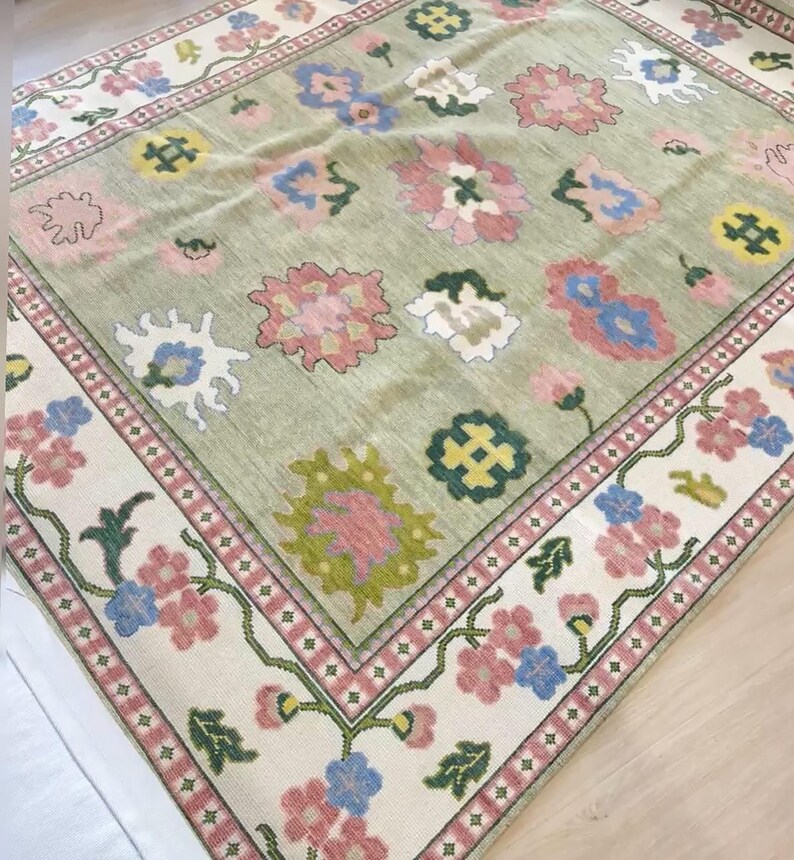 May include: A rectangular rug with a light green central field and a cream border. The rug features floral patterns in pink, blue, and yellow. The border has a repeating floral design with pink and green accents. The rug's dimensions are not visible.