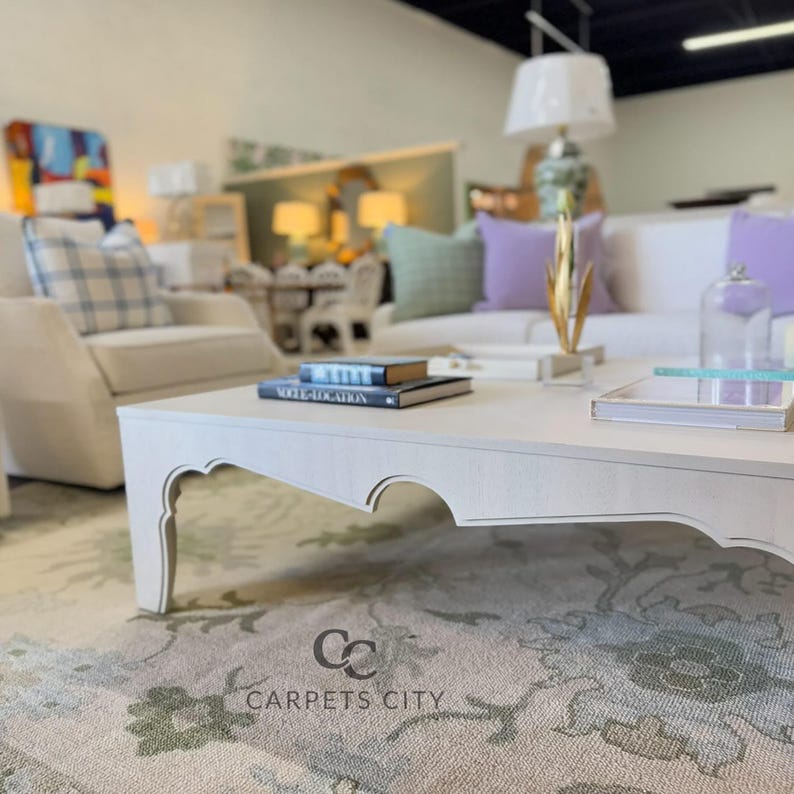 May include: A light-colored, ornate coffee table with a decorative edge, holding books and a tray. The table sits on a floral patterned rug. A white sofa with purple and green pillows is in the background. The logo for Carpets City is visible.