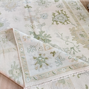 May include: Cream-colored area rug with a floral pattern in shades of green and beige. The rug has a folded corner, revealing the detailed border and fringe. The design features stylized botanical elements, creating a soft, inviting aesthetic.
