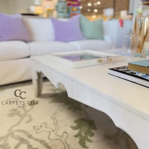 May include: A white coffee table with a scalloped edge, holding a tray and books. A white sofa with purple and green pillows is in the background. The logo for Carpets City is in the lower left corner.
