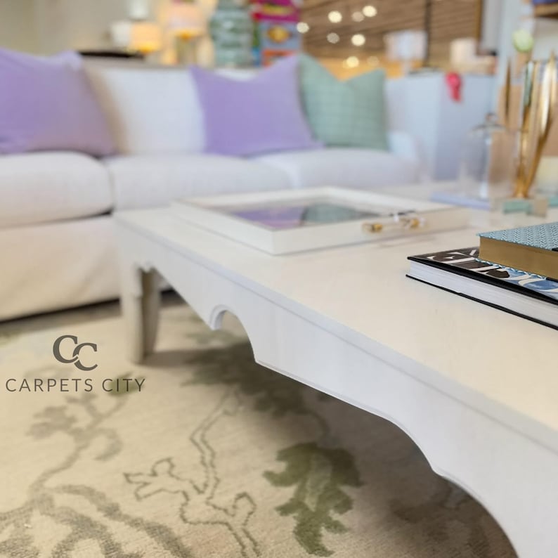 May include: A white coffee table with a scalloped edge, holding a tray and books. A white sofa with purple and green pillows is in the background. The logo for Carpets City is in the lower left corner.