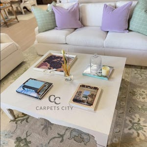 May include: A white square coffee table with decorative items, including books, a tray, and a glass cloche. The table has the text "CARPETS CITY" on the side. A white sofa with purple and green pillows is in the background.