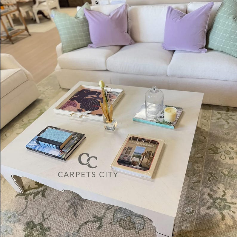 May include: A white square coffee table with decorative items, including books, a tray, and a glass cloche. The table has the text "CARPETS CITY" on the side. A white sofa with purple and green pillows is in the background.