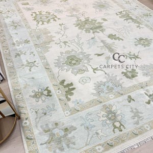 May include: A large, rectangular area rug with a floral pattern in shades of green, gray, and blue on a cream background. The rug has a detailed border and the text "CARPETS CITY" is visible. The rug is likely made of a soft material.