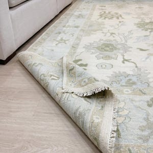May include: A cream-colored area rug with a floral pattern in shades of green and blue. The rug is partially rolled up, revealing a fringed edge. The rug is placed on a light wood floor.