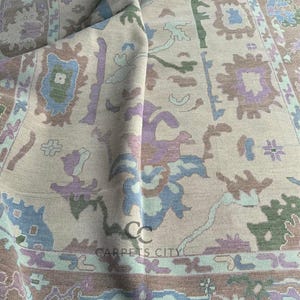 K&ouml;nnte beinhalten: Beiger Teppich mit einem detaillierten Muster in Blau-, Gr&uuml;n-, Lila- und Braunt&ouml;nen. Der Teppich hat ein zentrales Design mit floralen und geometrischen Elementen und einen Rand mit sich wiederholenden Motiven. Der Teppich ist gefaltet und zeigt die Textur und das Design.