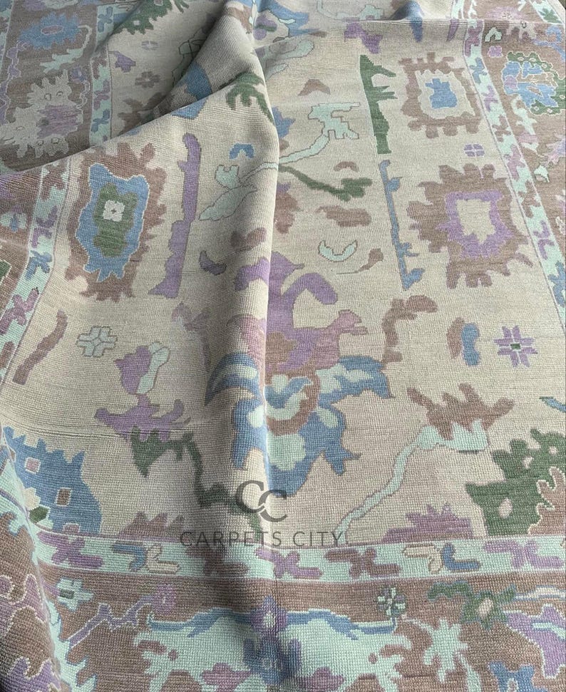 K&ouml;nnte beinhalten: Beiger Teppich mit einem detaillierten Muster in Blau-, Gr&uuml;n-, Lila- und Braunt&ouml;nen. Der Teppich hat ein zentrales Design mit floralen und geometrischen Elementen und einen Rand mit sich wiederholenden Motiven. Der Teppich ist gefaltet und zeigt die Textur und das Design.