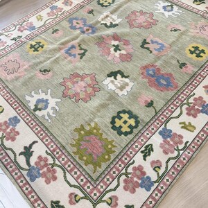 May include: A rectangular rug featuring a light green central field adorned with floral motifs in pink, blue, and yellow. The cream-coloured border showcases a repeating floral pattern. The rug has a traditional, vintage style.