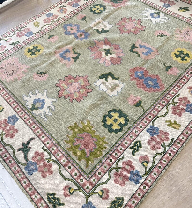 May include: A rectangular rug featuring a light green central field adorned with floral motifs in pink, blue, and yellow. The cream-coloured border showcases a repeating floral pattern. The rug has a traditional, vintage style.
