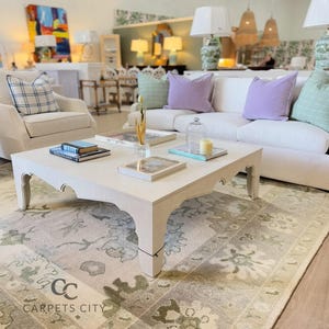 May include: A living room scene featuring a white square coffee table with decorative legs, a white sofa with pastel pillows, and a patterned rug. Books, a tray, and decorative items adorn the table. The room is well-lit, showcasing a blend of furniture and decor.
