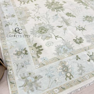 May include: A close-up of a cream-colored area rug with a floral pattern in shades of green, gray, and blue. The rug has a border with similar floral designs. The text "CARPETS CITY" is visible in the lower left corner.