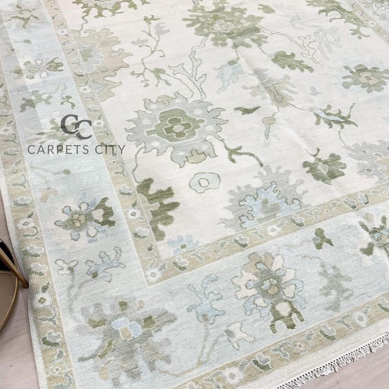 May include: A close-up of a cream-colored area rug with a floral pattern in shades of green, gray, and blue. The rug has a border with similar floral designs. The text "CARPETS CITY" is visible in the lower left corner.