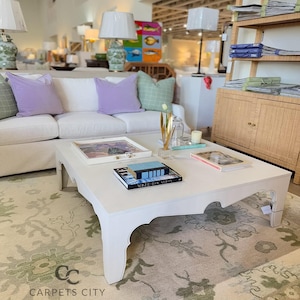 May include: A white square coffee table with curved edges, holding books, a tray, and a glass vase. A white sofa with lavender and green pillows sits behind the table. The room has a rug with a floral pattern.