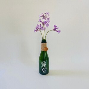 May include: A green glass bottle with a white floral design and a brown button tied around the neck. The bottle is filled with purple flowers.