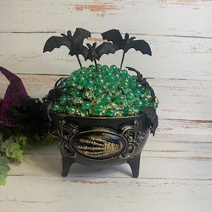 May include: A black cauldron with three legs and a gold skeleton hand emblem on the front. The cauldron is filled with shiny green beads and topped with three black bat figures and two black spider figures attached to the rim.
