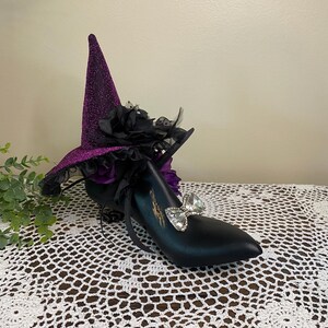 May include: A black shoe form adorned with a purple glittery pointed hat featuring black ruffled fabric, dark floral accents, and a feather. A clear, faceted, bow-shaped crystal embellishment is attached to the front of the black shoe form.