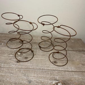 May include: Two rusty metal springs with circular and spiral designs. The springs are made of thin, dark brown metal and sit on a light-colored wooden surface. The springs have a vintage, industrial aesthetic.