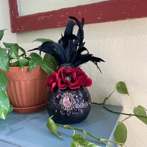 May include: Black and red Halloween-themed gourd with a black feather headdress and a decorative mirror. The gourd is decorated with gold paint and has a red rose and other floral accents.
