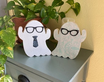 May include: Two white ghost figurines with black accents. One ghost wears a black tie and black glasses. The other ghost wears black glasses. The ghosts are sitting on a light gray wooden surface.