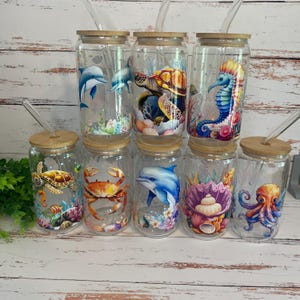 May include: A collection of clear glass tumblers with bamboo lids and glass straws. Each tumbler features a different colorful sea creature design, including dolphins, turtles, seahorses, crabs, and octopuses. The tumblers are designed for cold beverages.