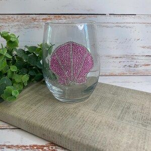 May include: A clear stemless wine glass featuring a decorative seashell design on the front. The seashell is composed of numerous small, bright pink rhinestones that create a textured, sparkling surface against the smooth glass exterior.