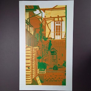 May include: A vertical linocut print depicting a courtyard scene with timber-framed architecture, a balcony, and stacked wooden crates. The artwork uses a palette of orange, yellow, green, and white, featuring a signature in the bottom right corner.