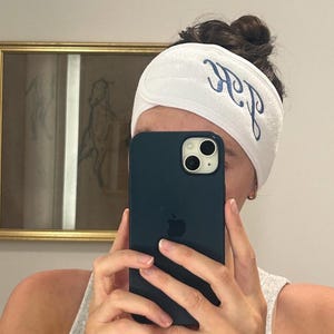 Op de afbeelding: Witte spa-hoofdband met blauw geborduurde initialen. De hoofdband is gemaakt van een zacht, absorberend materiaal en is ontworpen om haar uit het gezicht te houden. De persoon houdt een donkerblauwe smartphone vast.