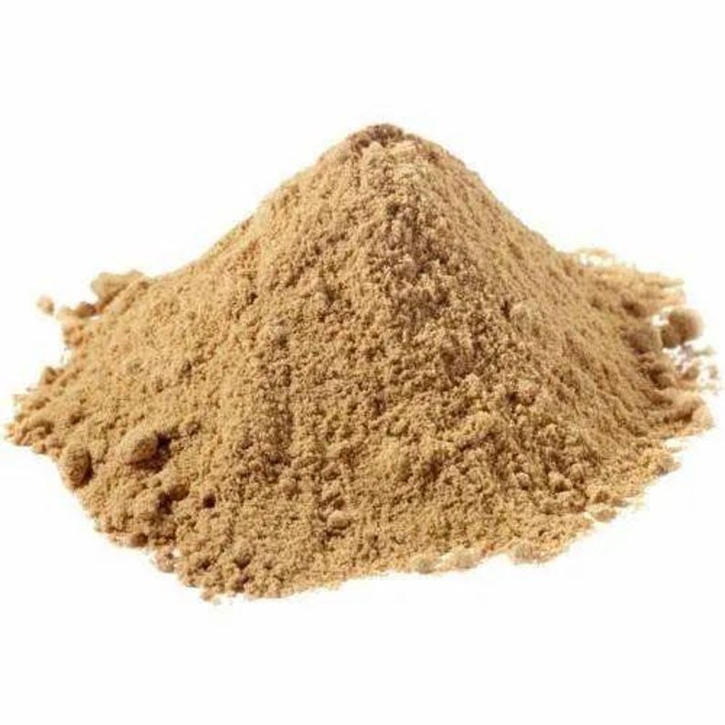 May include: A pile of light brown powder against a white background. The powder has a fine, granular texture and is slightly darker in the center, creating a sense of depth. The overall shape is a rounded mound.