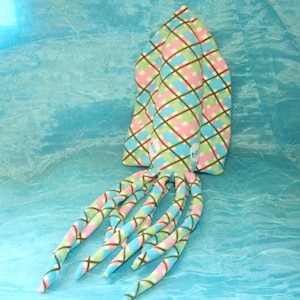 May include: A plush, patterned squid toy in shades of pink, blue, green, and brown. The squid has a diamond pattern on its head and tentacles. The background is a light blue, textured surface.