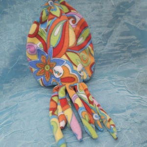 May include: A colorful plush octopus toy with a psychedelic floral pattern in orange, red, blue, and yellow. The toy has eight tentacle-like appendages and white button eyes. The background is a light blue fabric.