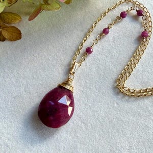 May include: A gold chain necklace featuring a large, faceted, teardrop-shaped deep pink gemstone pendant wrapped in gold wire. The chain is accented with small, round, deep pink beads spaced along the links, creating a delicate design.