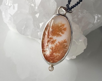 May include: A silver pendant necklace with a white oval agate stone featuring a dendritic pattern in shades of brown. The pendant is attached to a black chain.