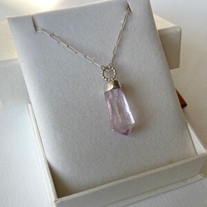 Puede incluir: Un collar de cadena de plata con un colgante de cristal rosa p&aacute;lido en una caja blanca. El cristal est&aacute; facetado y tiene una punta puntiaguda.